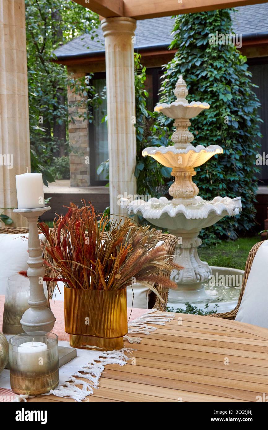 Elegante fontana in pietra in un'accogliente terrazza giardino, circondata da sedie in vimini, vegetazione e arredamento rustico. Un tranquillo ambiente esterno con arco classico Foto Stock