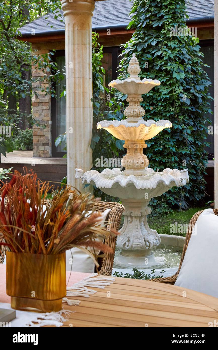 Elegante fontana in pietra in un'accogliente terrazza giardino, circondata da sedie in vimini, vegetazione e arredamento rustico. Un tranquillo ambiente esterno con arco classico Foto Stock