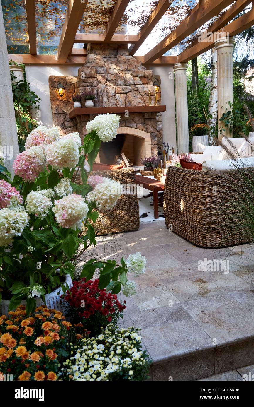 Accogliente terrazza all'aperto con caminetto in pietra, sedie in vimini e fiori colorati in fiore. Un elegante patio con giardino che mescola fascino rustico e naturale Foto Stock