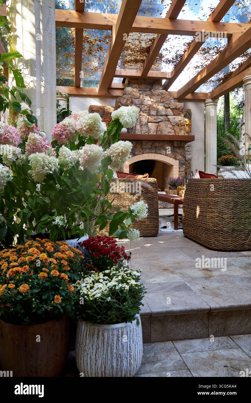 Accogliente terrazza all'aperto con caminetto in pietra, sedie in vimini e fiori colorati in fiore. Un elegante patio con giardino che mescola fascino rustico e naturale Foto Stock
