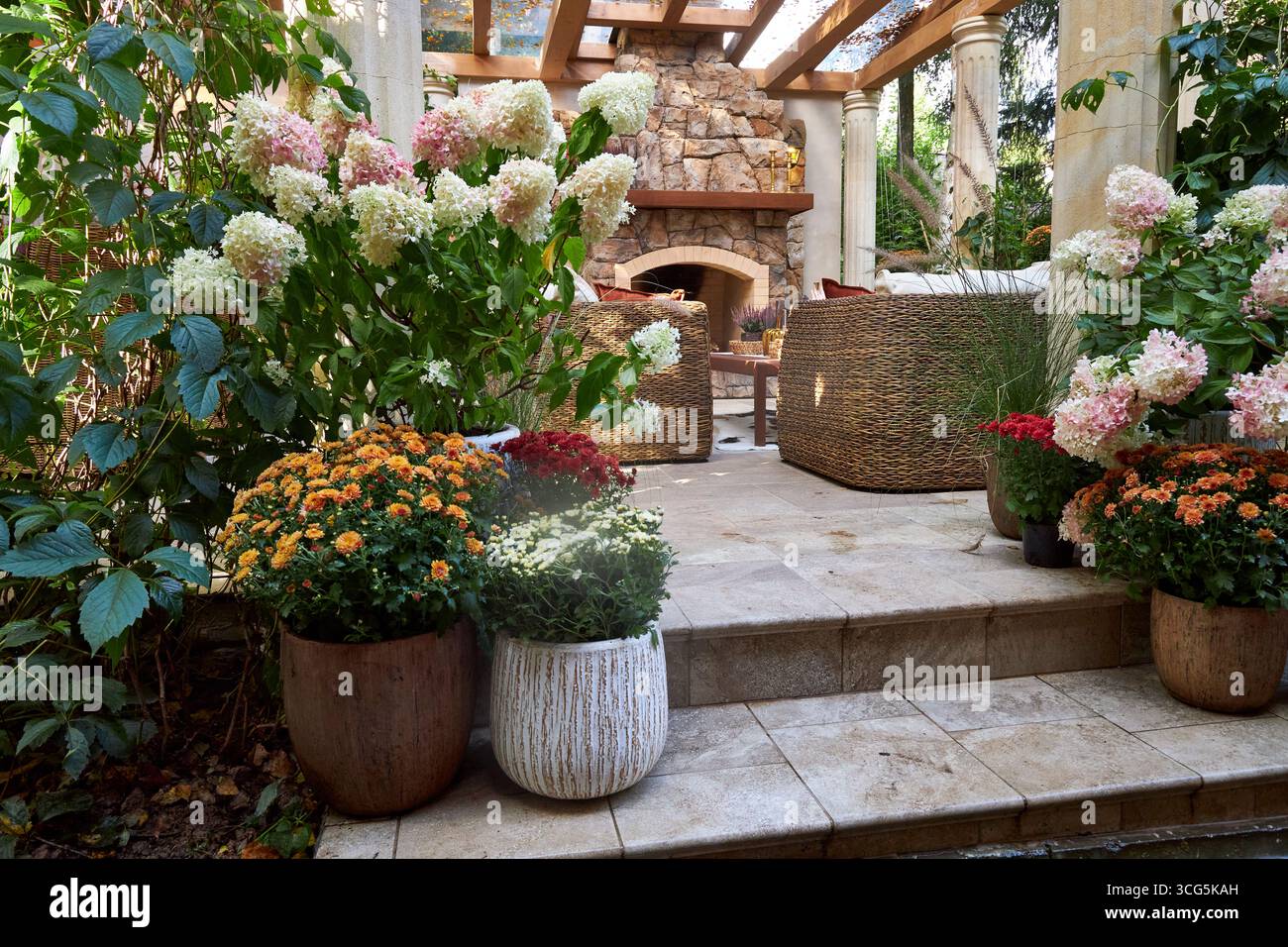 Accogliente terrazza all'aperto con caminetto in pietra, sedie in vimini e fiori colorati in fiore. Un elegante patio con giardino che mescola fascino rustico e naturale Foto Stock