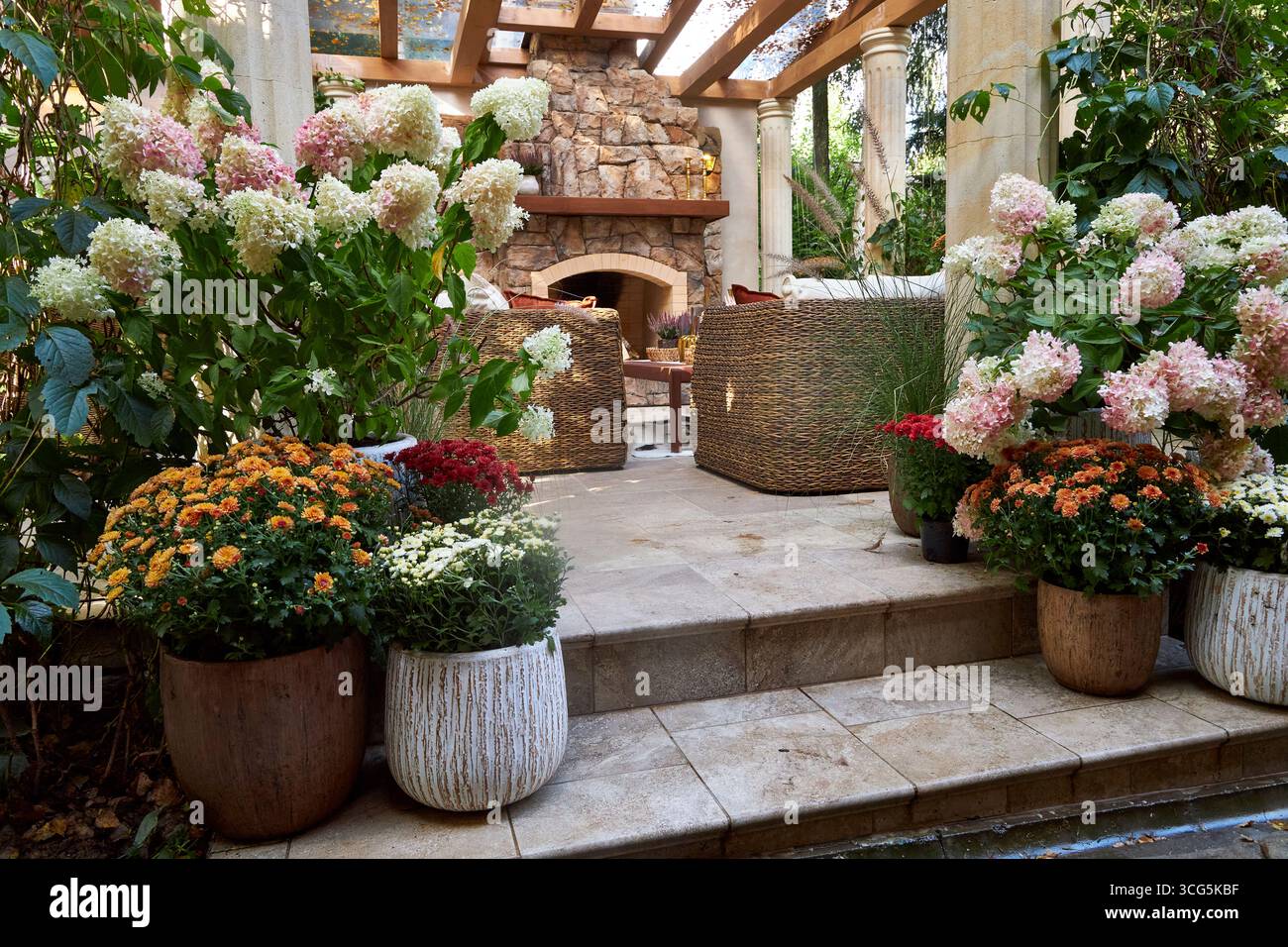 Accogliente terrazza all'aperto con caminetto in pietra, sedie in vimini e fiori colorati in fiore. Un elegante patio con giardino che mescola fascino rustico e naturale Foto Stock