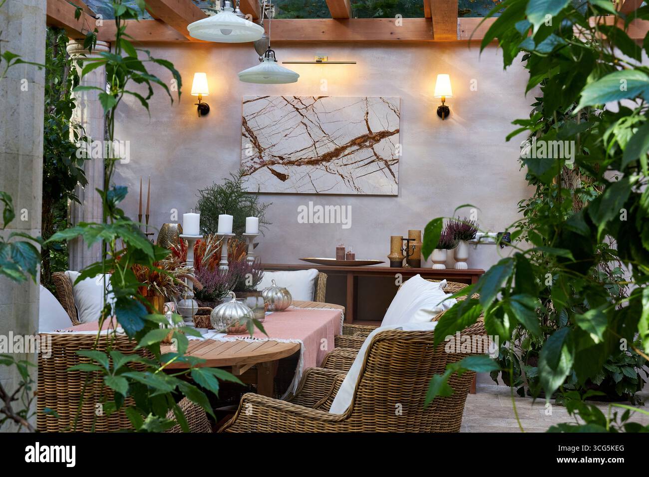 Elegante zona pranzo all'aperto con sedie in vimini, tavolo in legno, candele e piante decorative. Le luci da parete calde creano un'atmosfera accogliente e completa Foto Stock