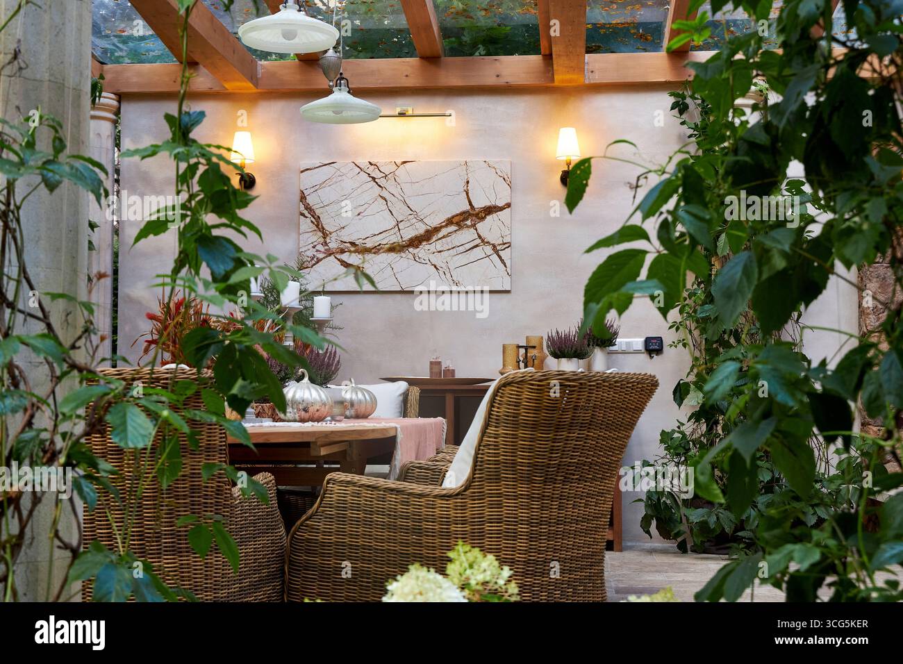 Elegante zona pranzo all'aperto con sedie in vimini, tavolo in legno, candele e piante decorative. Le luci da parete calde creano un'atmosfera accogliente e completa Foto Stock