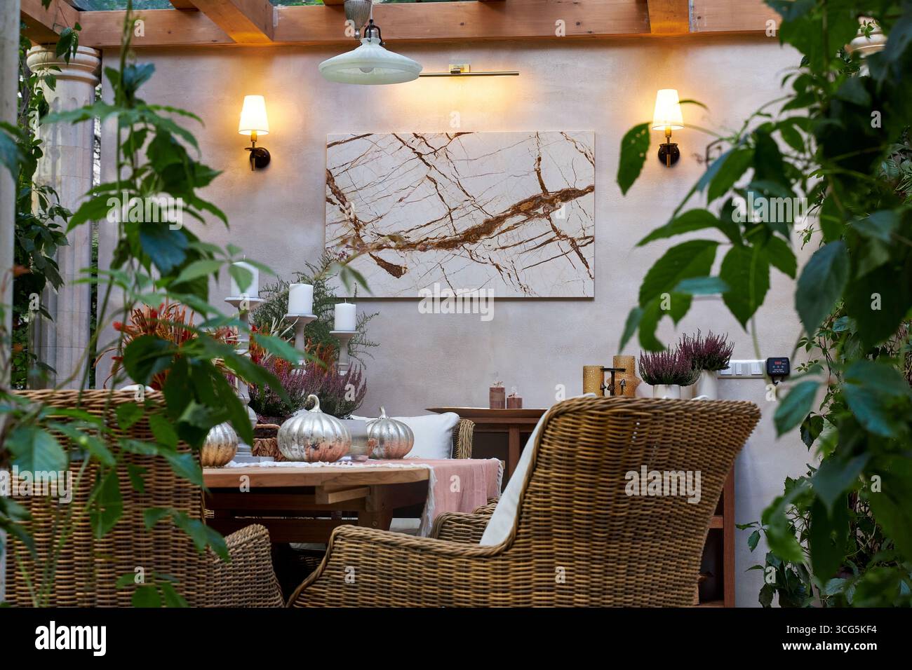 Elegante zona pranzo all'aperto con sedie in vimini, tavolo in legno, candele e piante decorative. Le luci da parete calde creano un'atmosfera accogliente e completa Foto Stock
