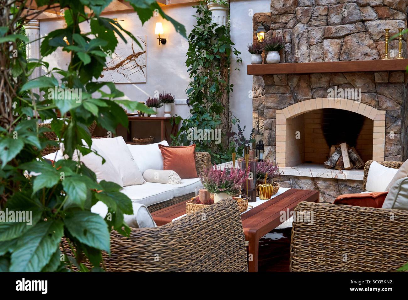 Accogliente area soggiorno all'aperto con caminetto in pietra, sedie in vimini, cuscini morbidi e luce naturale calda. Elegante design rustico combinato con vegetazione Foto Stock