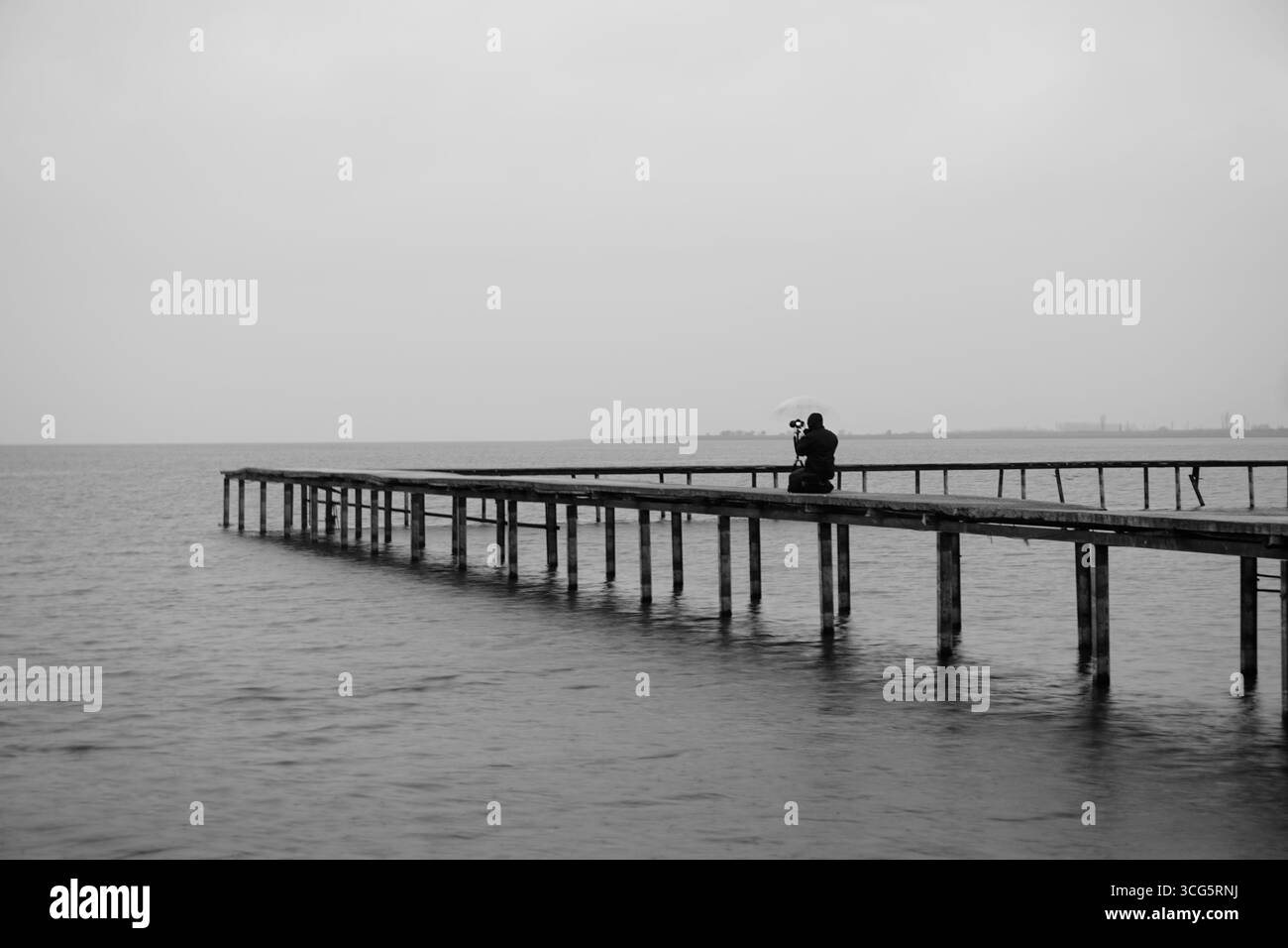 Fotografia in bianco e nero di una persona su un molo desolato con un ombrello, di fronte al mare grigio Foto Stock