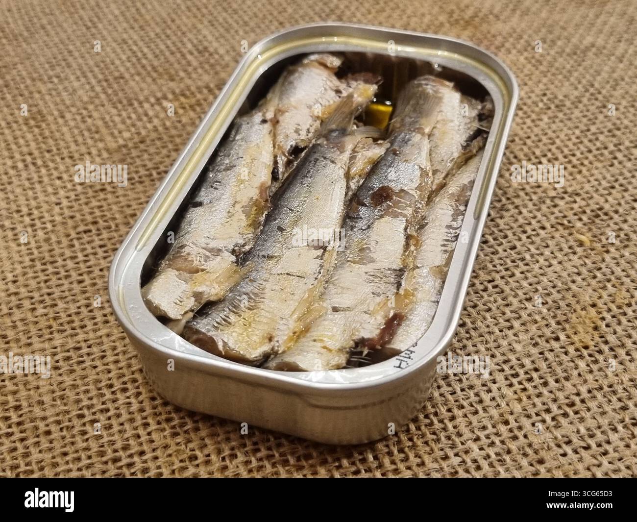 Una lattina aperta di sardine posta su tessuto di bavone; le sardine sono allineate in modo ordinato, evidenziando la consistenza argentata, l'olio e il contenitore metallico. Foto Stock