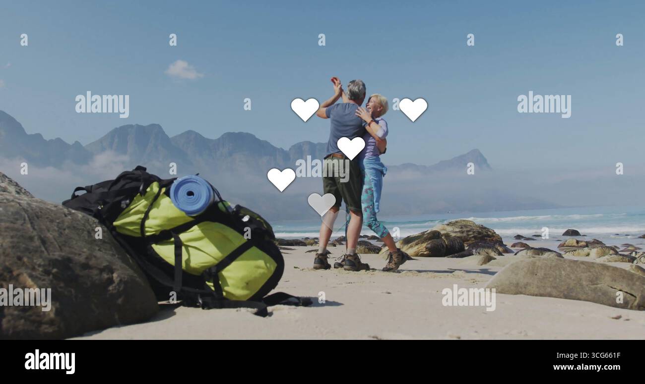 Coppia danzante che indossa abiti da trekking che si muove sulla riva sabbiosa, con zaino giallo e massi Foto Stock