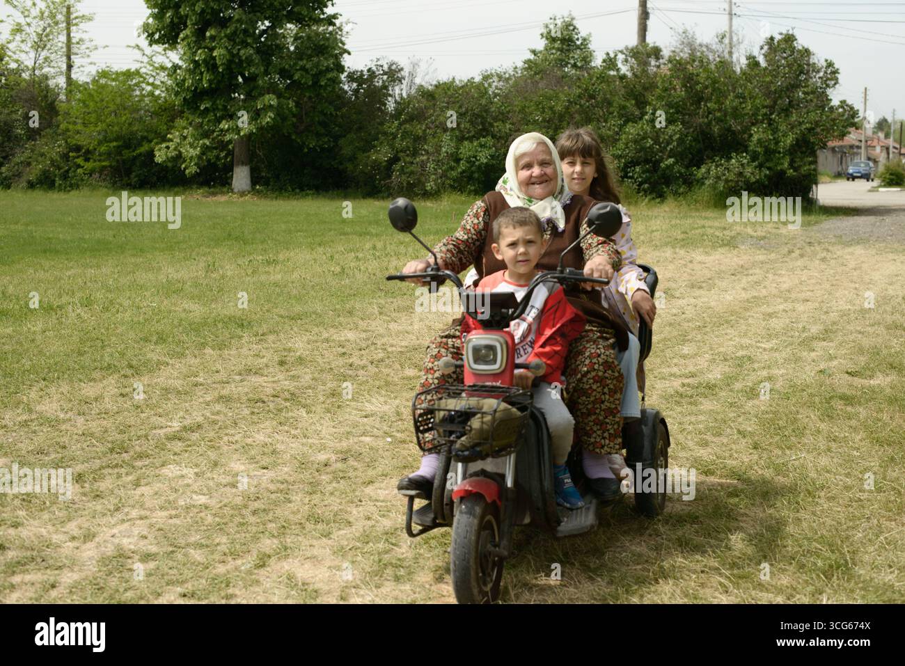 Una nonna Alevi a Sevar, Bulgaria, guida un triciclo elettrico con i suoi nipoti, riflettendo la mobilità rurale, le tradizioni familiari e intergen Foto Stock