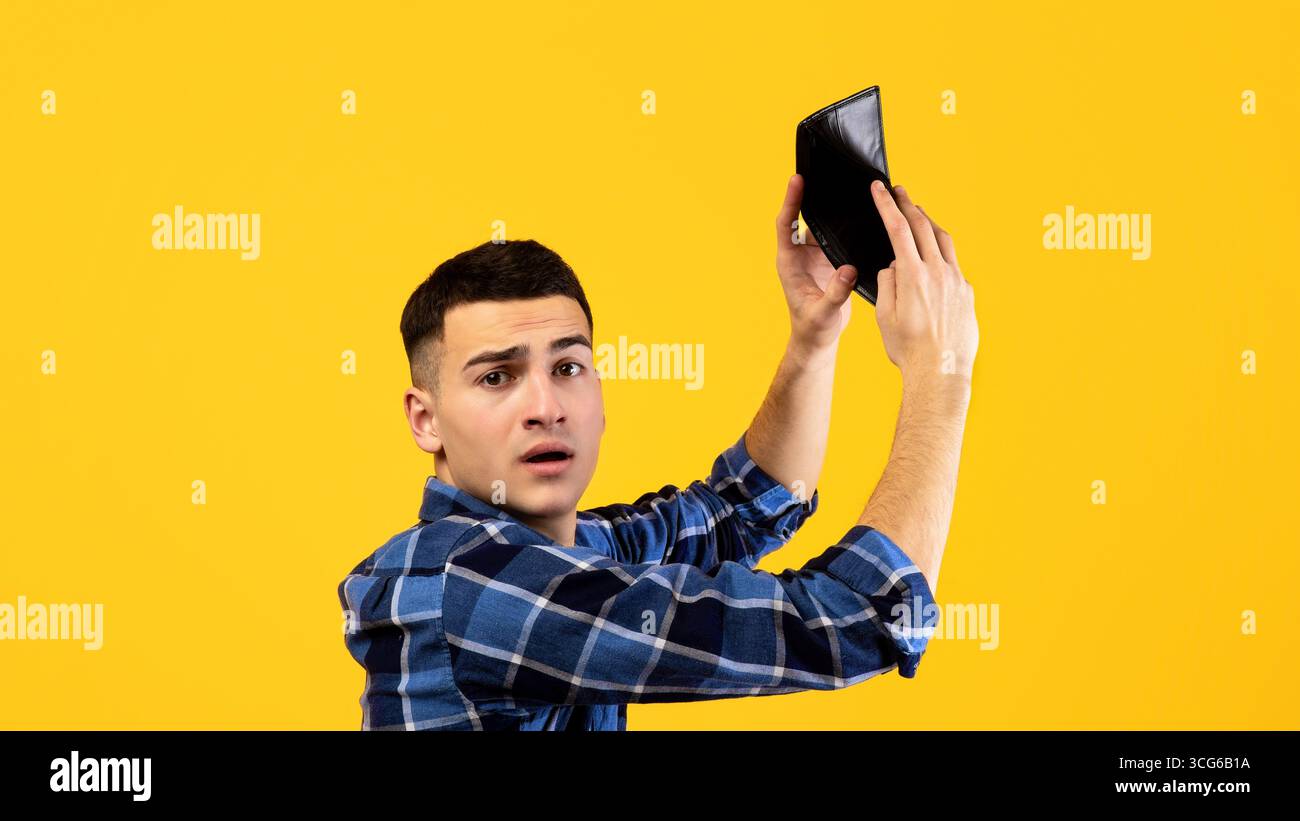 Uomo millenario infelice scuotendo portafoglio vuoto su sfondo studio arancione Foto Stock