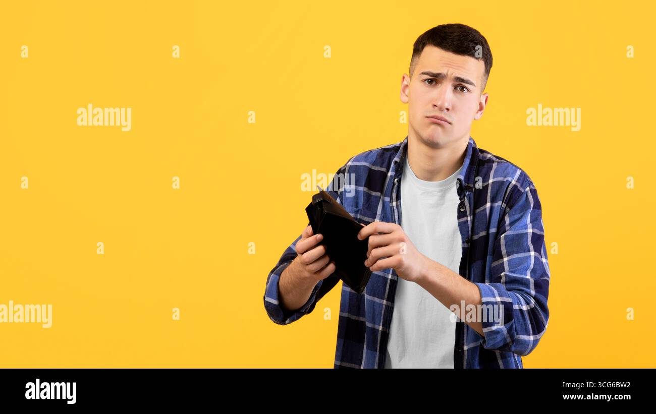 Concetto di fallimento. Un giovane uomo sconvolto che mostra un portafoglio vuoto, che soffre di perdita di denaro su sfondo studio arancione Foto Stock