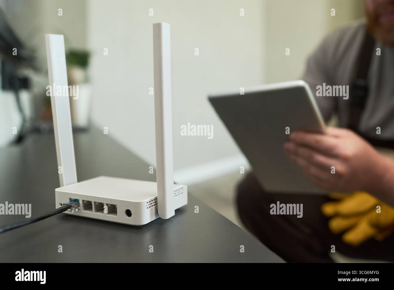 Uomo di mezza età caucasico che tiene un tablet digitale seduto vicino al router wireless sul tavolo, concentrandosi sul processo di configurazione del dispositivo in un moderno ambiente domestico o di ufficio Foto Stock