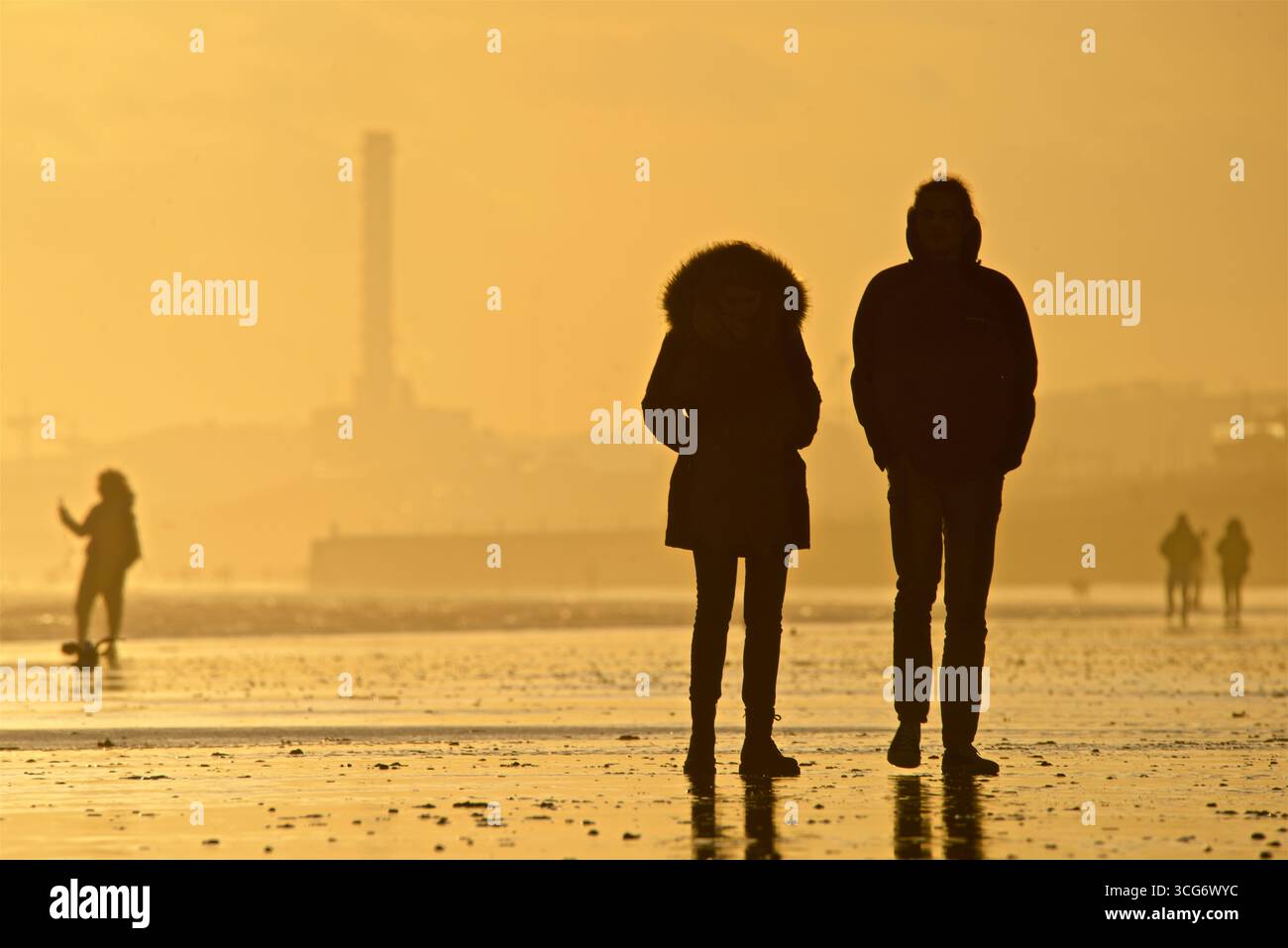 Coppia che si gode la bassa marea al tramonto. Sagome di persone e Shoreham Power Station in lontananza. Brighton e Hove, Inghilterra, Regno Unito Foto Stock