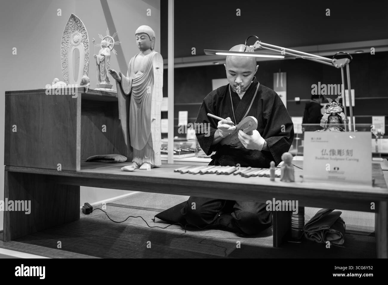 Un artigiano crea una scultura di Buddha in legno in uno studio tradizionale, che riflette l'artigianato culturale. Gli attrezzi e i pezzi finiti possono essere osservati su Foto Stock Un artigiano crea una scultura di Buddha in legno in uno studio tradizionale, che riflette l'artigianato culturale. Gli attrezzi e i pezzi finiti possono essere osservati su Foto Stock