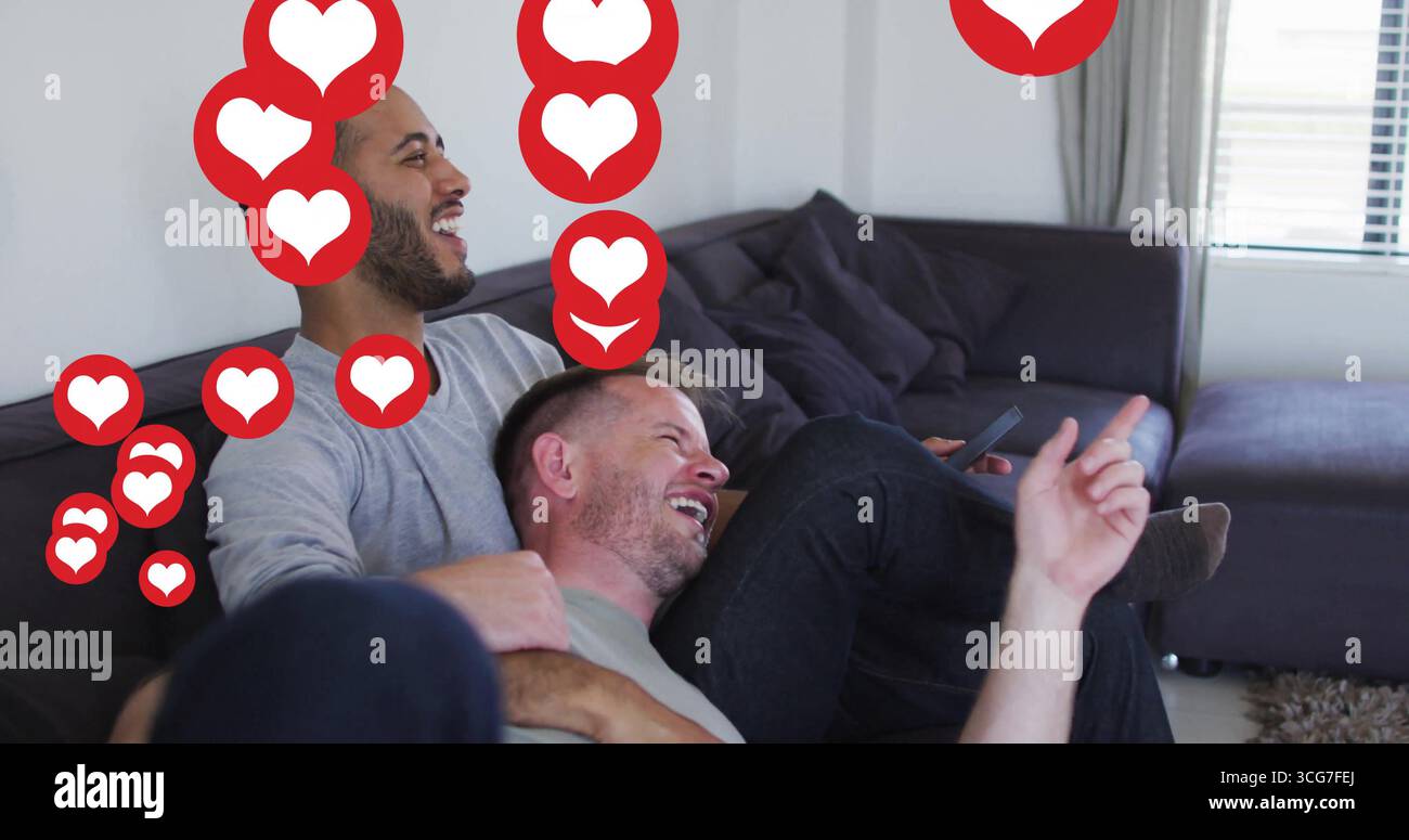 Uomini che ridono di mezza età seduti su un divano sezionale grigio nel soggiorno, con smartphone e icone del cuore Foto Stock