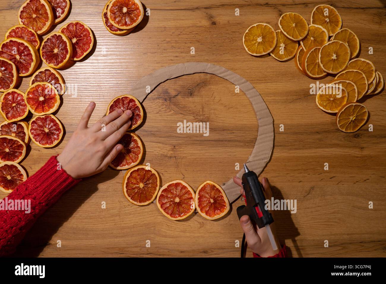 Donna che utilizza colla calda corona di Natale fatta a mano da fette di pompelmo d'arancia secca. Preparazione per le festività natalizie, maestria nella creazione creativa dell'arredamento. Attività pratiche creazione di un workshop di regali per le festività sostenibili e responsabili. Vista dall'alto, disposizione piatta Foto Stock