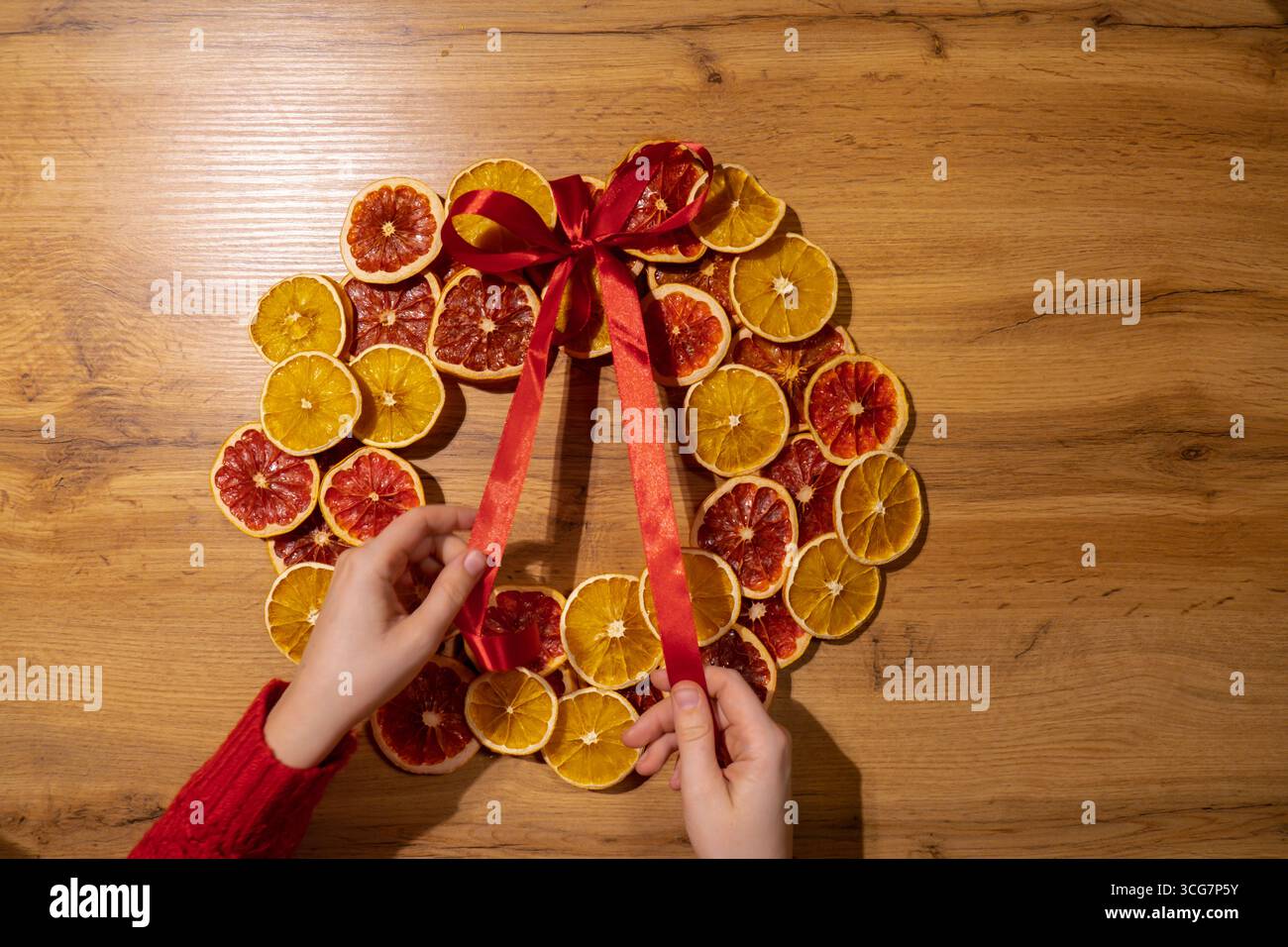 Fase 3 nastro di legatura fatto a mano corona di Natale da fette di pompelmo d'arancia secca. Preparazione per le festività natalizie, maestria nella creazione creativa dell'arredamento. Attività pratiche creazione di un workshop di regali per le festività sostenibili e responsabili. Vista dall'alto, disposizione piatta Foto Stock