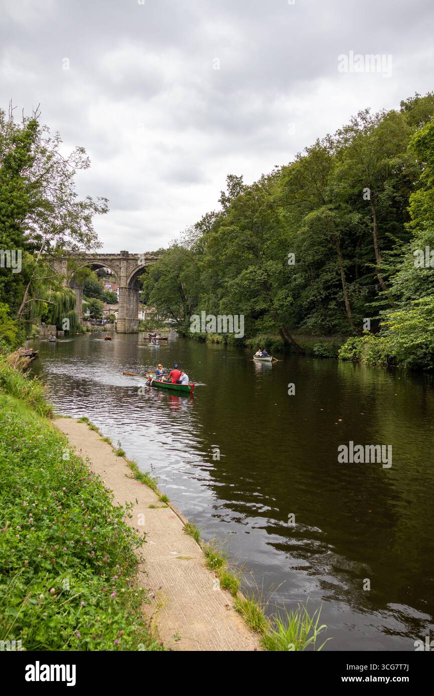 Una piccola barca a remi con persone su un fiume tranquillo circondato da fitti alberi verdi, con un vecchio ponte in pietra in lontananza sotto un cielo nuvoloso Foto Stock