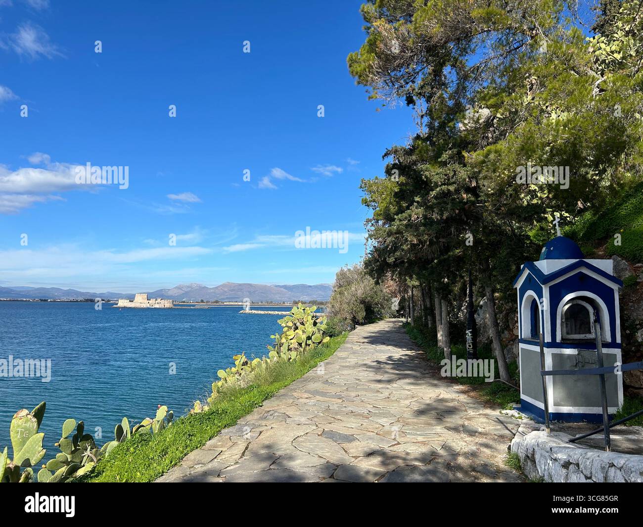 Passeggiata costiera con un piccolo santuario lungo la strada e vista della fortezza di Bourtzi a Nafplio, Peloponneso, Grecia. - Immagine stock catturata con smartphone
