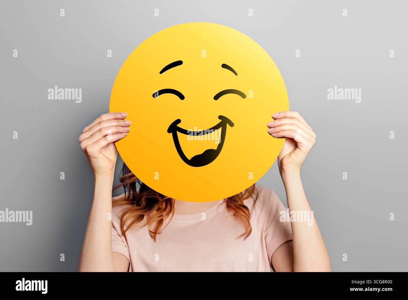 Felice emoji. La ragazza tiene un'emoticon gialla con un sorriso isolato su uno sfondo grigio. Felice emoji Foto Stock