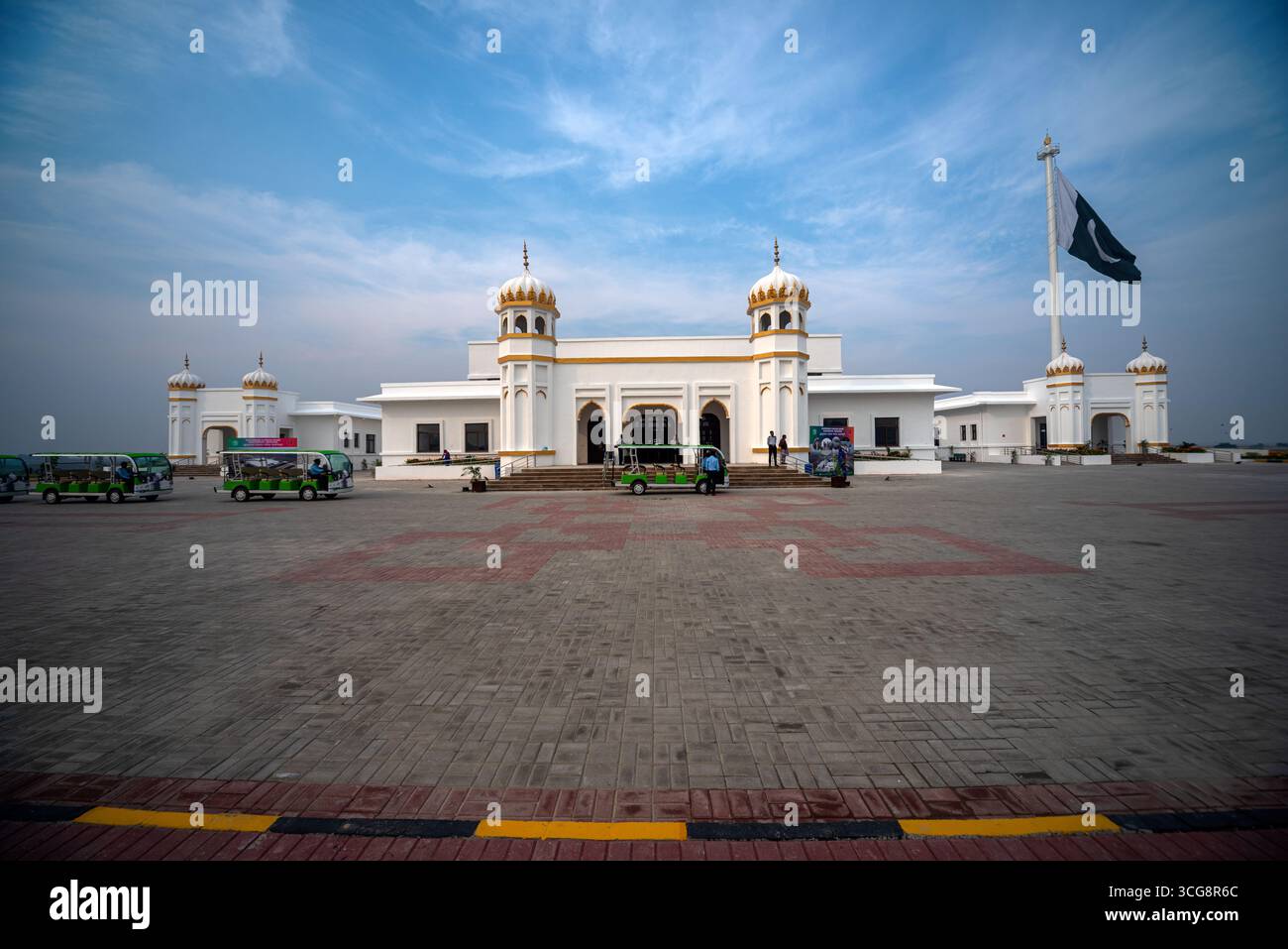 Vista di un edificio bianco con cupole dorate sotto un cielo luminoso accanto a una bandiera ondulata, con veicoli colorati parcheggiati nelle vicinanze, Kartarpur, Punjab, Pakistan. Foto Stock