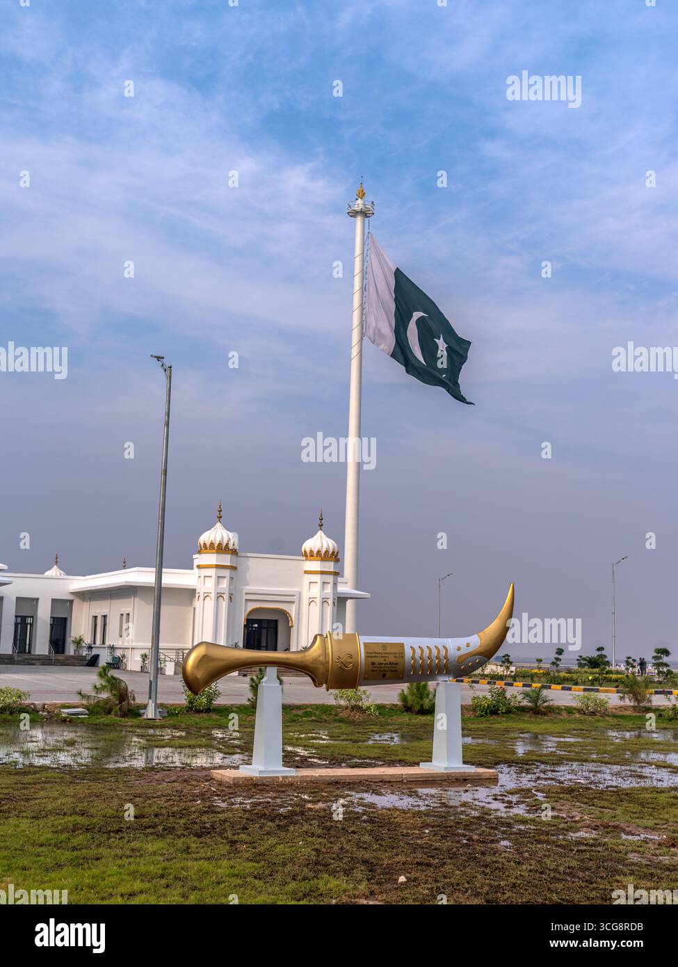 La vista di una statua di kirpan dorata risplende sotto la bandiera pakistana, sullo sfondo di un edificio bianco incontaminato, Kartarpur, Punjab, Pakistan. Foto Stock