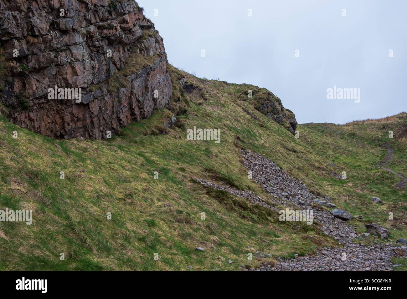 La vista di un'aspra scogliera incontra un ripido pendio erboso punteggiato di ghiaia rocciosa sotto un cielo nuvoloso, creando un contrasto di texture e colori, Cullen, Scozia, Regno Unito. Foto Stock