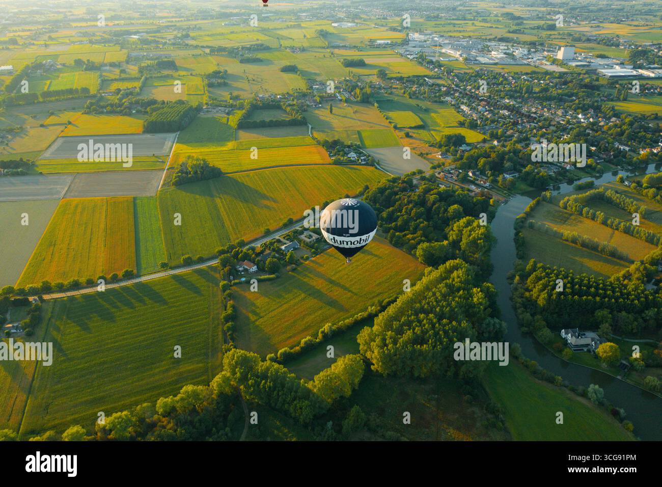 Sint-Martens-Latem, Belgio - 25 agosto 2025: Vista aerea di una mongolfiera che si sposta su campi verdeggianti e il tortuoso fiume Leie, un arazzo di verdi e gialli sotto la luce soffusa del mattino. Foto Stock
