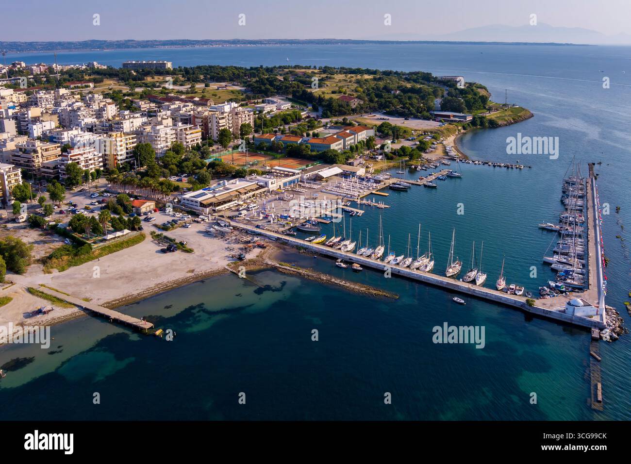 Vista aerea del lungomare orientale di Salonicco con porticciolo, edifici urbani, vegetazione lussureggiante e montagne lontane sotto un cielo limpido. Foto Stock