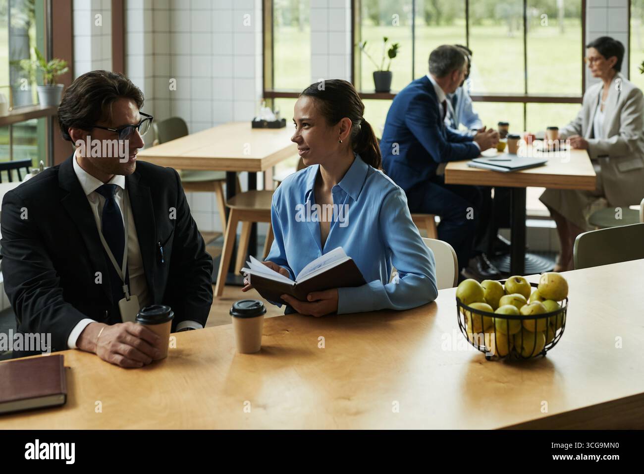 Giovane uomo e donna caucasica in abbigliamento da lavoro seduti a tavola tenendo tazze da caffè e libro, parlando in una moderna caffetteria con due colleghi di mezza età in sottofondo Foto Stock