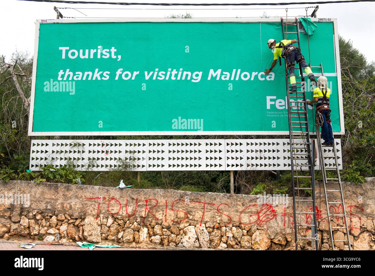 Arbeiter kleben ein Werbeplakat dass sich bei Touristen für dem Besuch Mallorcas bedankt. Darunter steht ein Graffiti Tourist torna a casa. Foto Stock