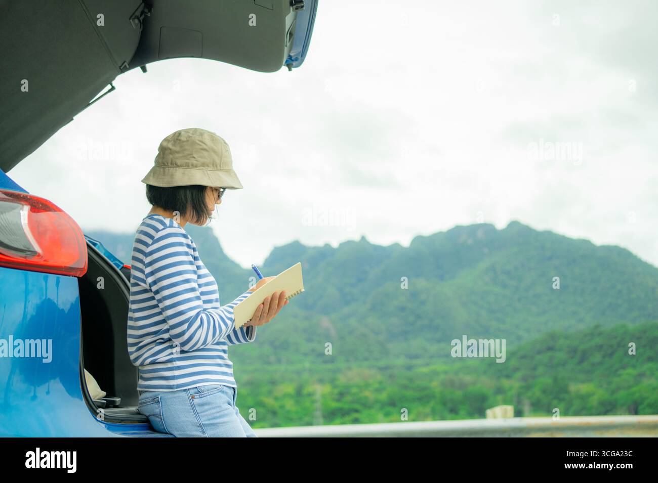 Donna che scrive su un notebook per la disintossicazione digitale. Riflesso di sé, abitudine giornalistica e pianificazione consapevole accanto a un'auto blu con paesaggio montano Foto Stock