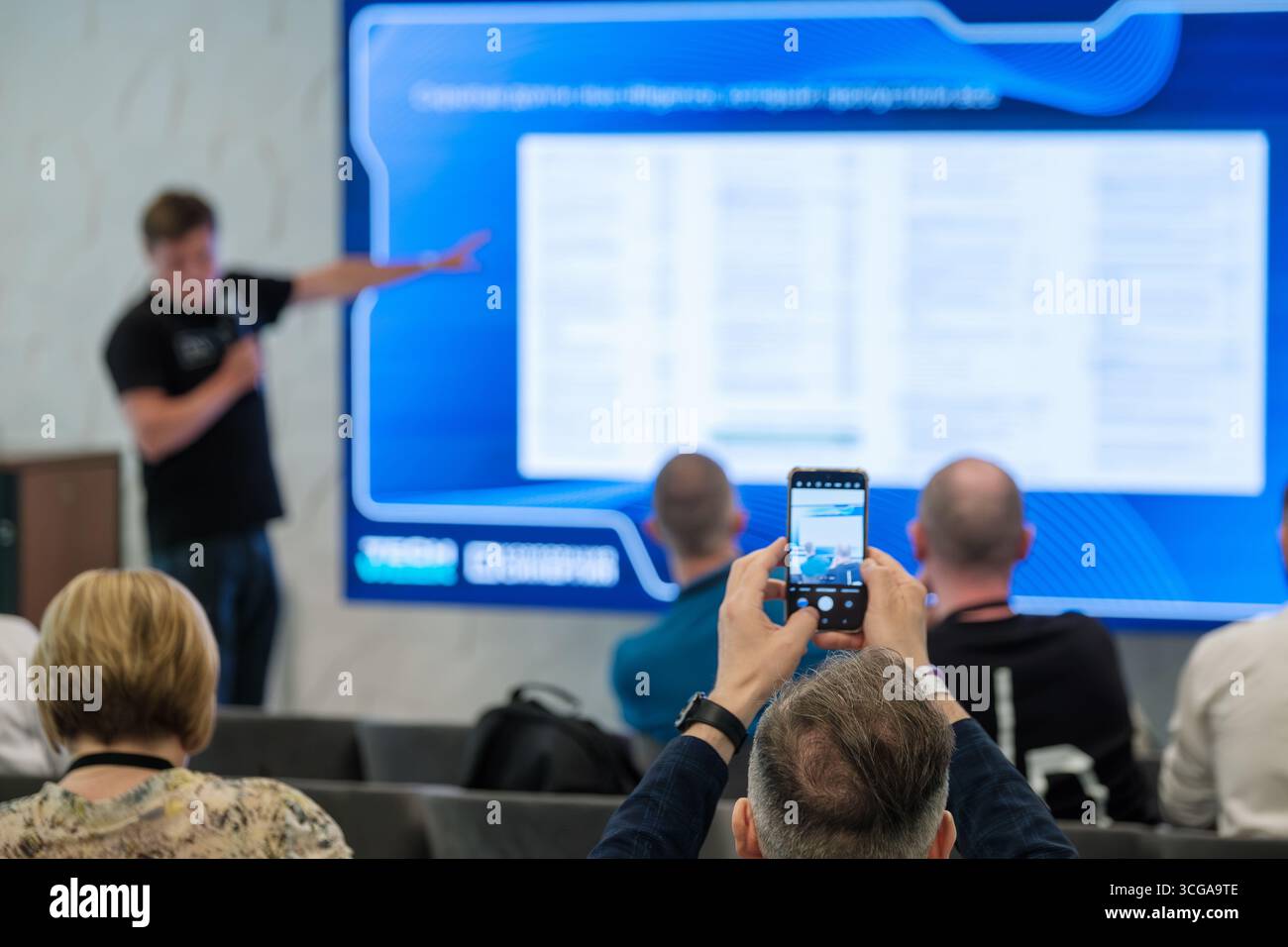 Partecipanti a un seminario al chiuso che registra una presentazione professionale sul proprio smartphone, sottolineando l'apprendimento visivo e le tecnologie moderne. Foto Stock