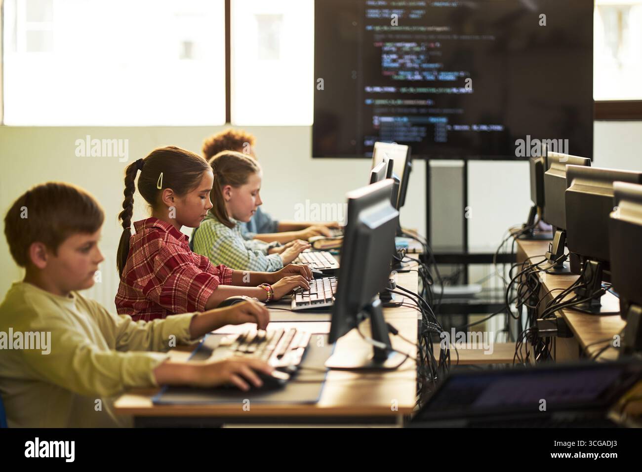 Gruppo di bambini caucasici e neri seduti alle scrivanie utilizzando computer desktop, digitando e concentrandosi sul codice di programmazione visualizzato su un grande monitor in una moderna aula Foto Stock