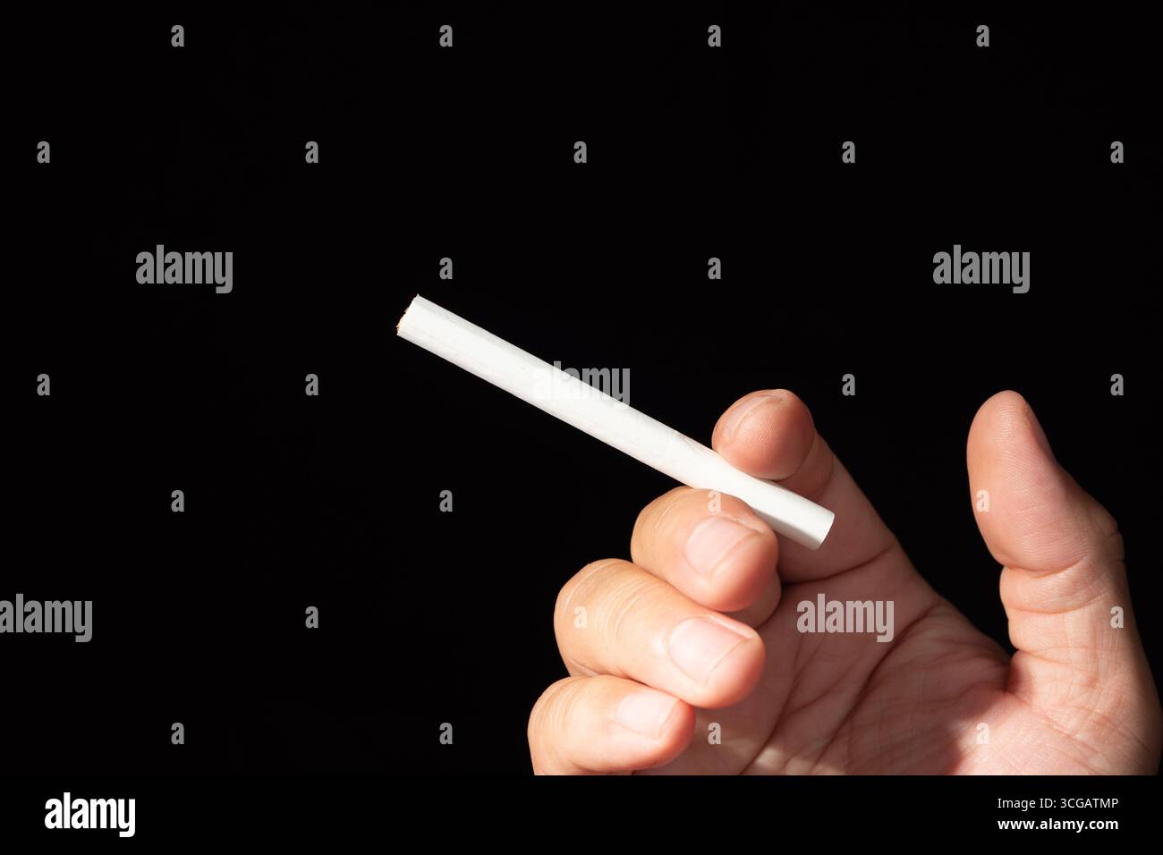 Una mano tiene una sigaretta accesa con fumo che si arriccia delicatamente verso l'alto su uno sfondo scuro e contrastante. Foto Stock