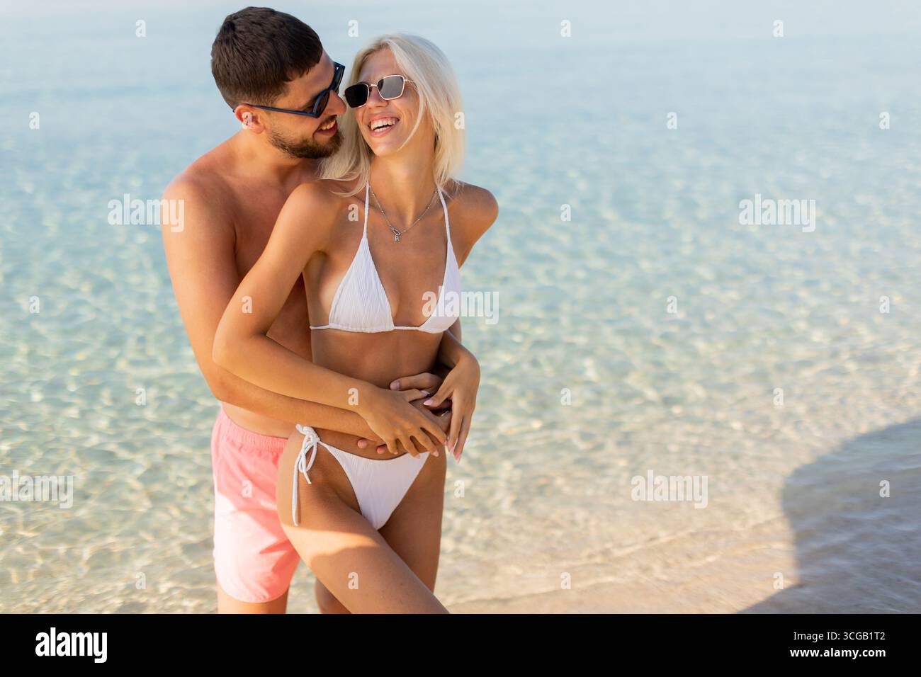 Due amanti si abbracciano gioiosamente lungo la costa, crogiolandosi nel calore del sole e in compagnia l'uno dell'altro, circondati da un mare tranquillo. Foto Stock
