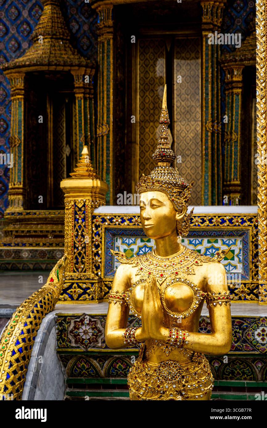 Una statua dorata di una figura femminile in posa di preghiera, adornata da intricati gioielli, adagiata su uno sfondo del tempio riccamente decorato con colorate t Foto Stock