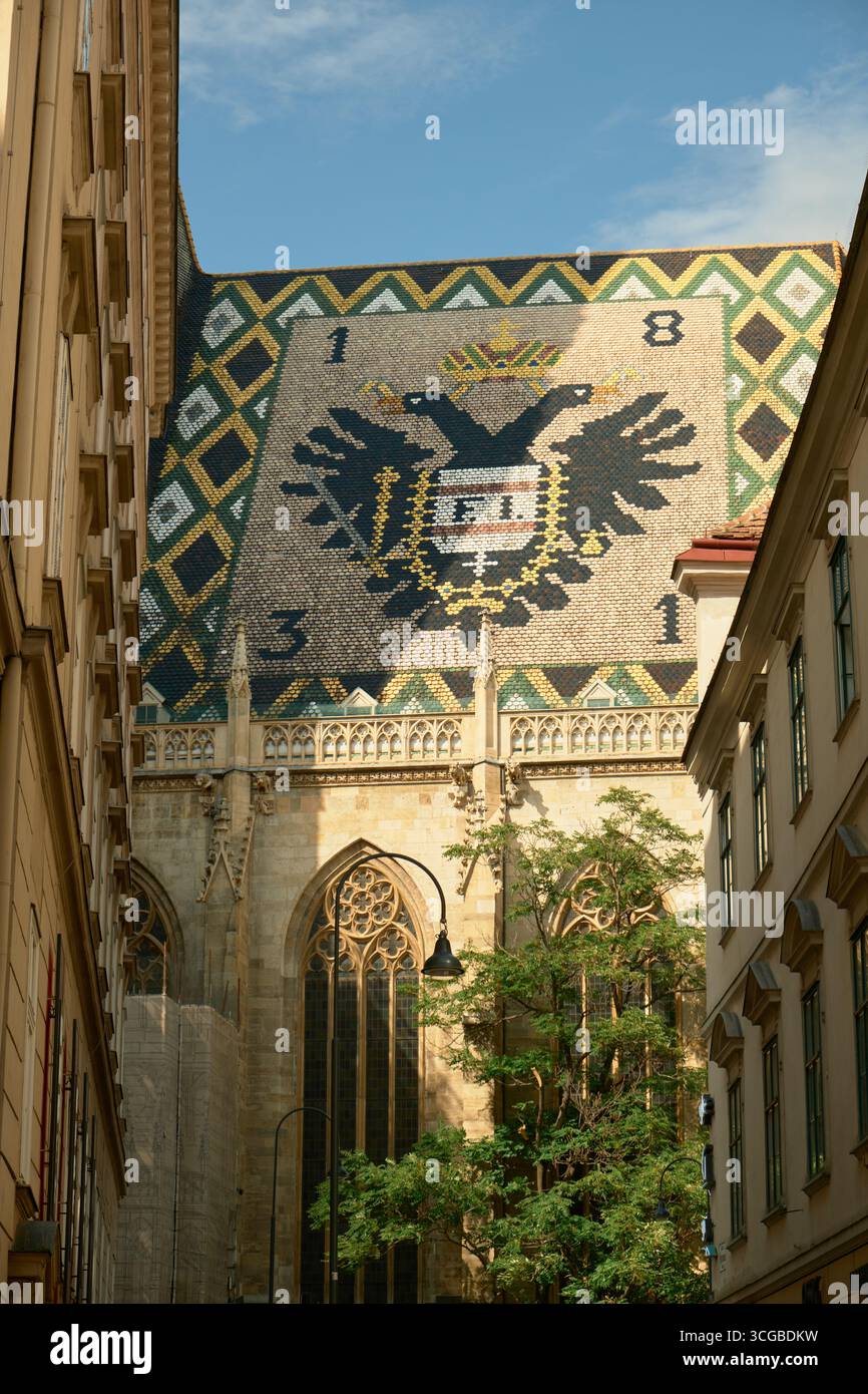 Primo piano della cattedrale di Santo Stefano a Vienna, con il suo tetto a mosaico illuminato dal sole con simboli dell'aquila imperiale, grandi vetrate colorate e un albero Foto Stock