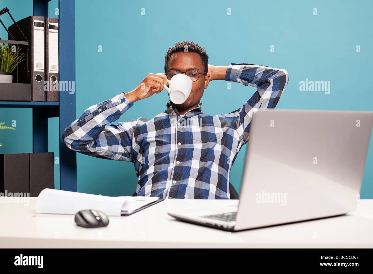Uomo afroamericano che si rilassa sulla sedia e beve un caffè mentre guarda un video sul suo portatile. Giovane seduto alla scrivania, appoggiato all'indietro con una tazza in mano e guardando lo schermo del dispositivo. Foto Stock