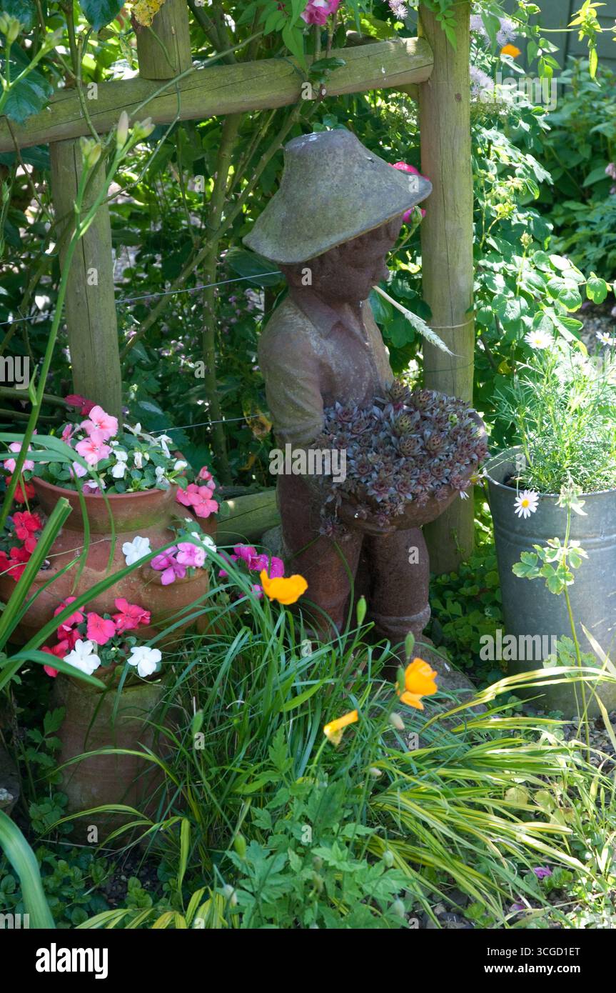 Una stravagante statua di un bambino si erge tra fiori colorati in un lussureggiante giardino pieno di vegetazione durante una soleggiata giornata estiva. Foto Stock