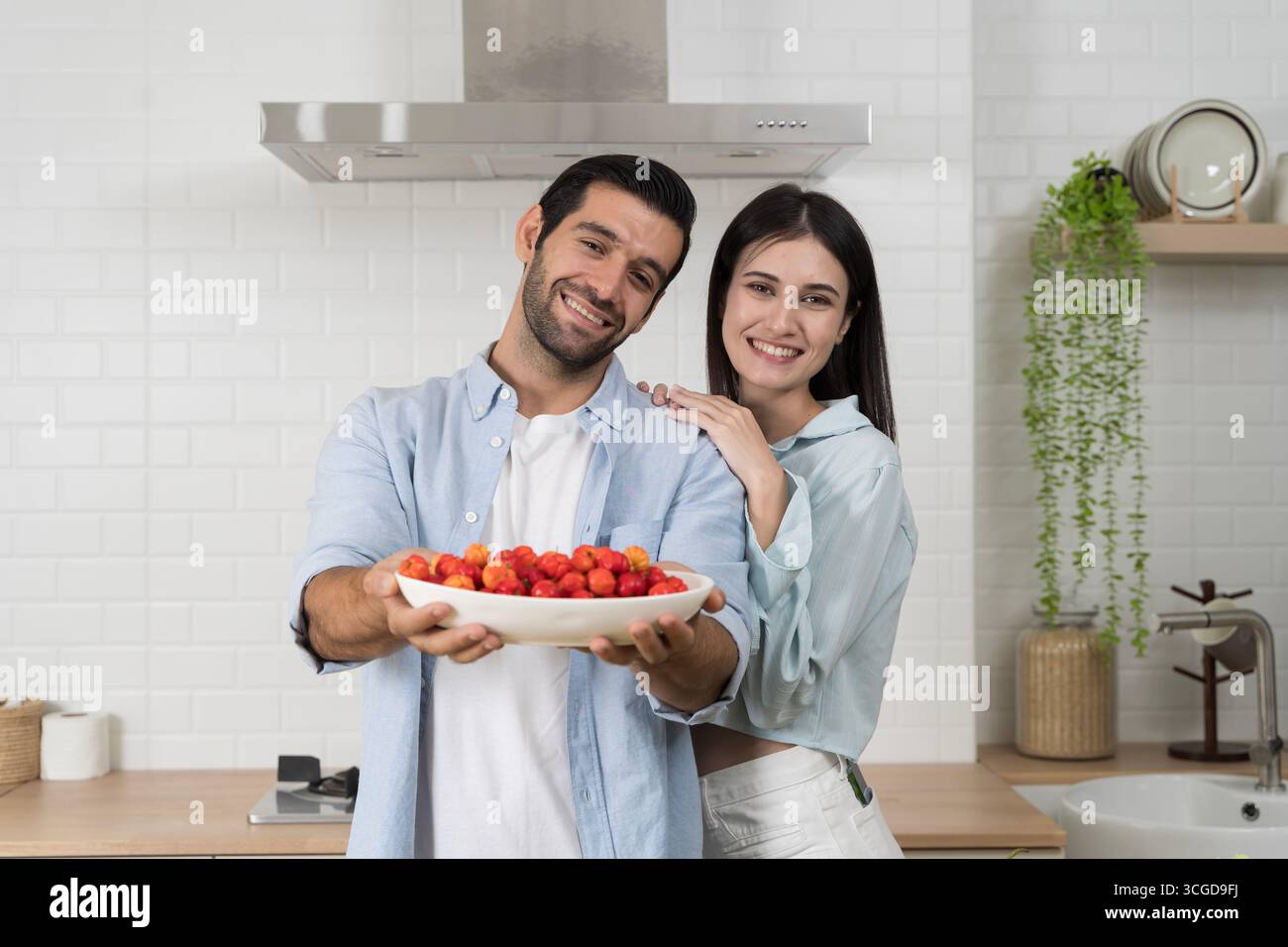 Coppia felice in una cucina moderna che mostra un piatto di ciliegie fresche, che rappresentano l'amore, il benessere, il cibo biologico e uno stile di vita sano, con un'emozione luminosa Foto Stock