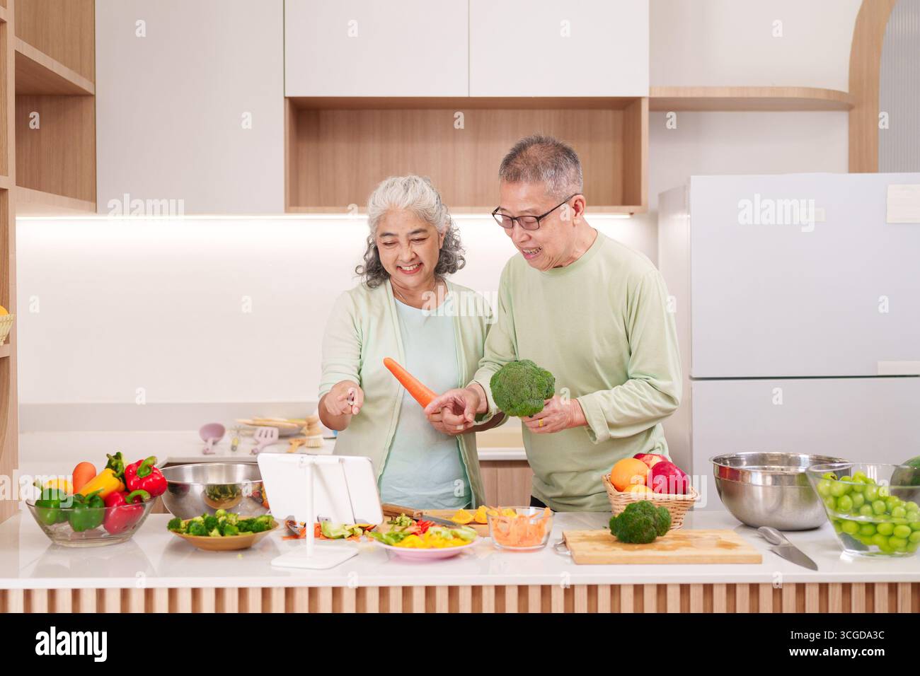 Felice coppia asiatica più anziana che cucina verdure fresche in una cucina moderna e luminosa. Seguono una ricetta digitale per un pasto sano. Godersi la vita domestica. Foto Stock