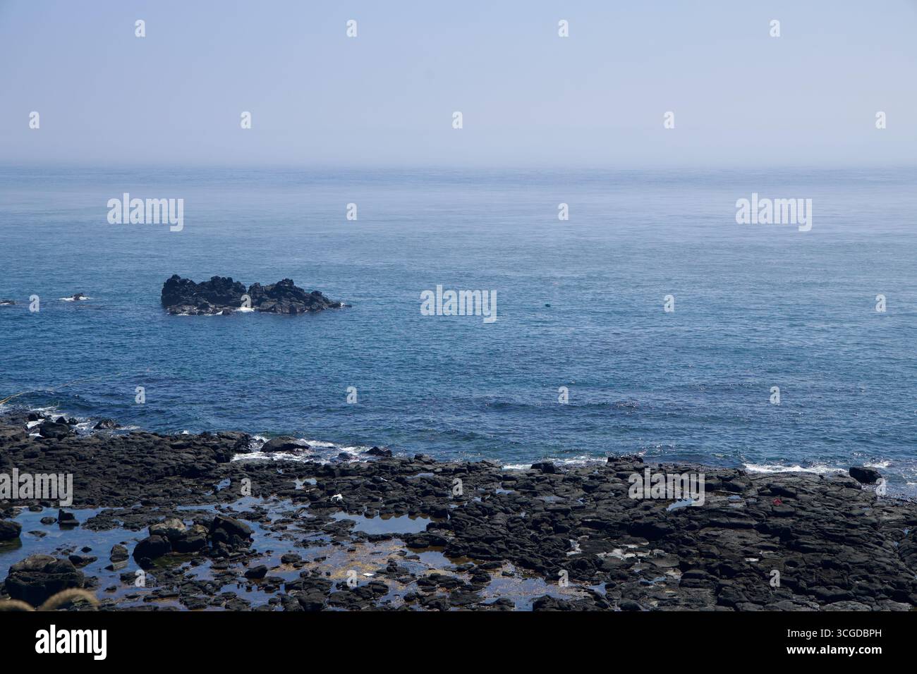 Un isolotto basalto scuro sorge appena al largo della costa occidentale di Jeju, circondato da calme acque blu e piscine di marea sparse attraverso la frastagliata piattaforma di lava u Foto Stock
