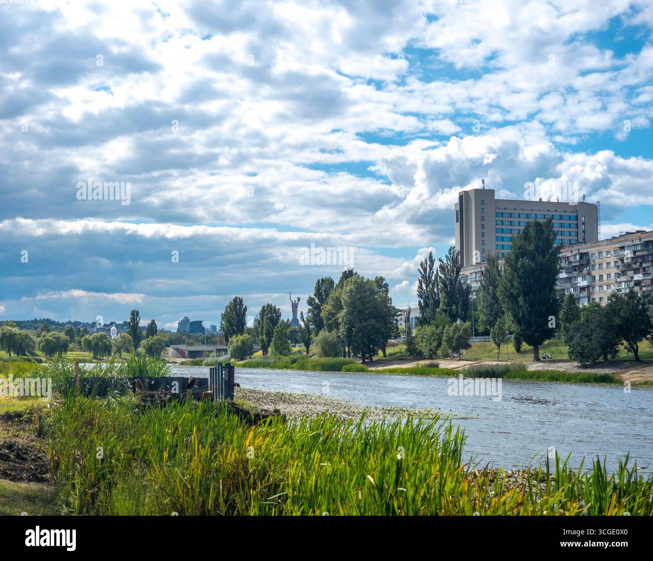 La lussureggiante vegetazione verde che costeggia il fiume Dnipro scorre pacificamente sotto un cielo nuvoloso, con l'iconico Monumento alla madre Ucraina che si innalza in lontananza, cre Foto Stock