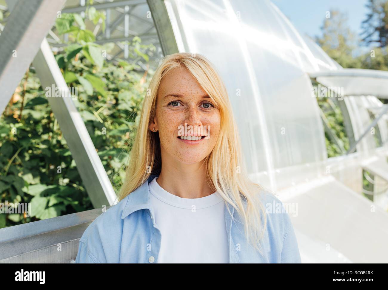 Donna sorridente con capelli biondi e lentiggini in piedi in una serra. Ritratto estremamente dettagliato di un lavoratore della serra in piedi all'aperto. Foto Stock