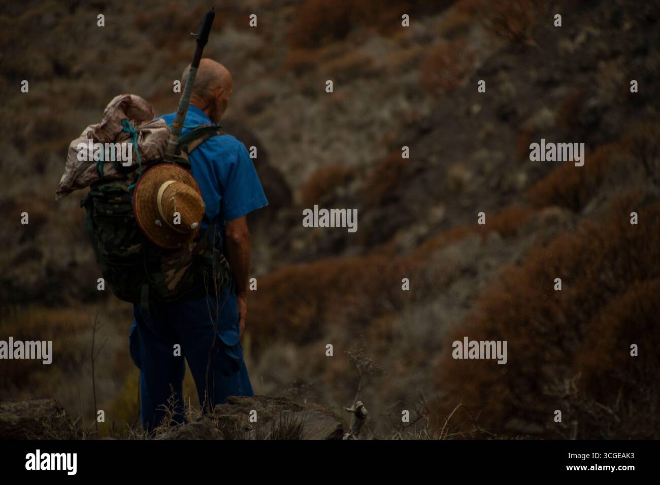 Vecchio pescatore con camicia blu e cappello di paglia si stende con zaino e canna da pesca, guardando le colline aspre, lo stile di vita di fatica e tradizione. Foto Stock