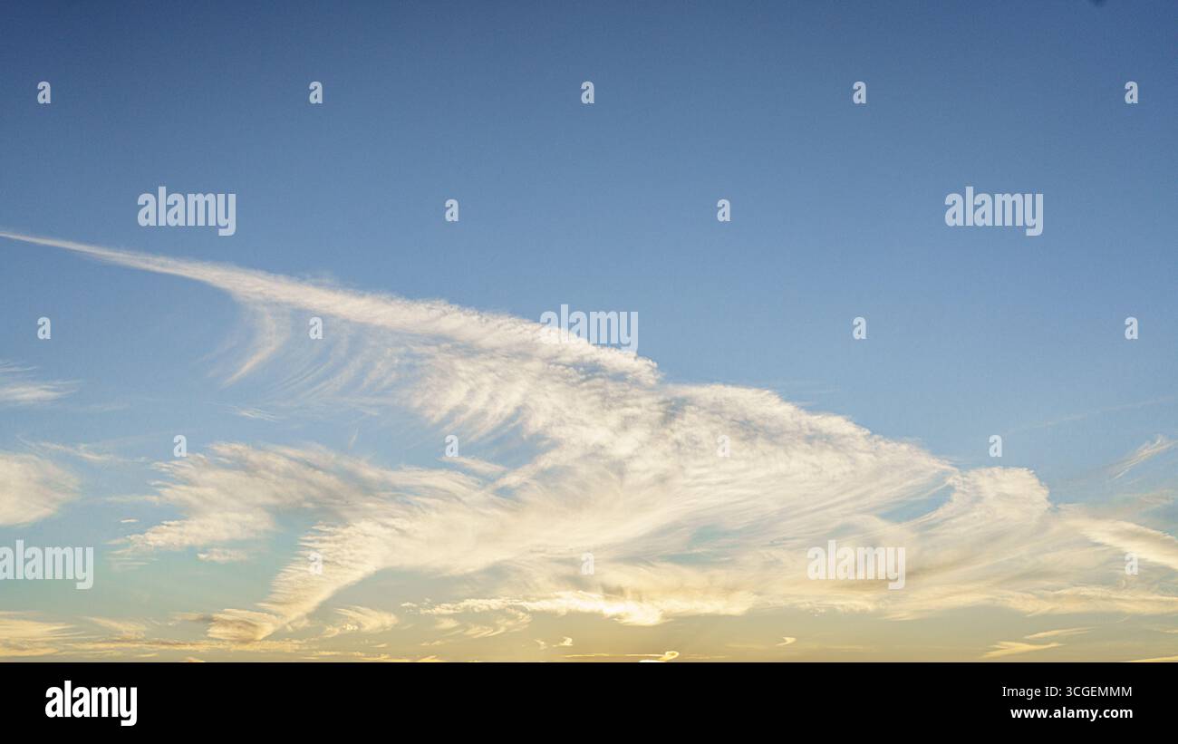 Le delicate nuvole di cirrus si estendono su un cielo blu durante l'ora d'oro, illuminate delicatamente dal sole. Foto Stock