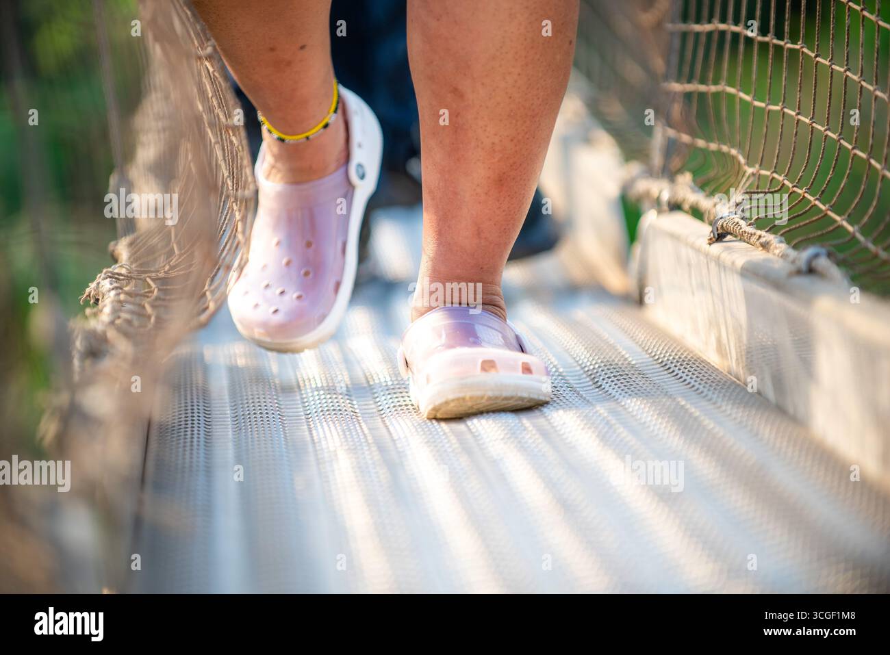 Primo piano delle gambe di una persona che indossano scarpe casual in stile Crocs mentre cammina con attenzione attraverso un ponte sospeso con lati a rete, a simboleggiare l'equilibrio Foto Stock