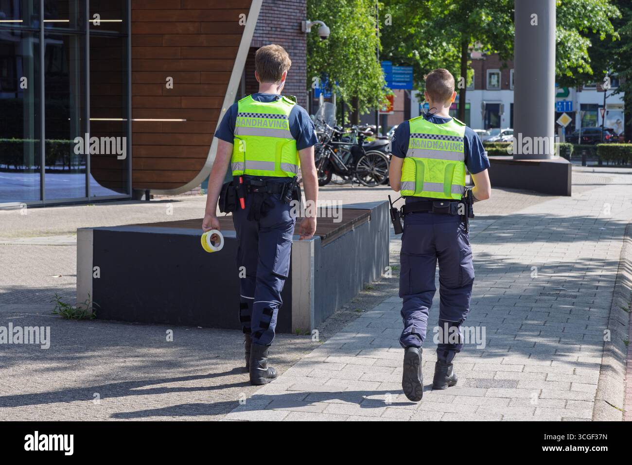Due agenti di "Handhaving" (forze dell'ordine) in giubbotti riflettenti si allontanano dalla telecamera su un'ampia strada asfaltata di Amersfoort. Amersfoort, Paesi Bassi, 1 Foto Stock